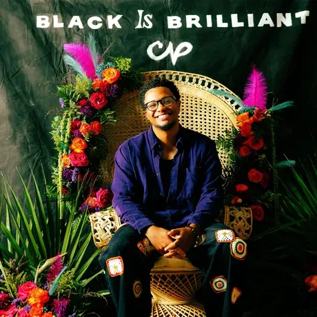 Adam Mahoney sits in a rattan chair decorated with tropical flowers and feathers. Neon sign behind him says "beaucoup power: Black is brilliant". Indeed it is.