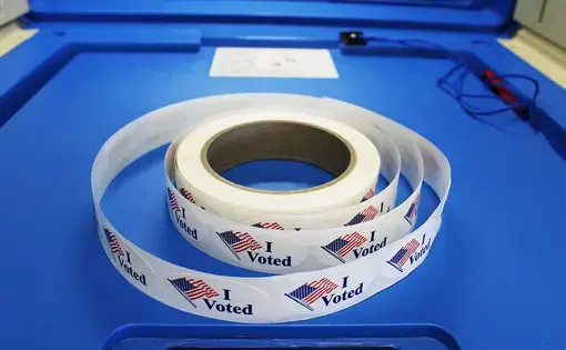 Red white and blue "I voted" stickers with American flag are on a spool of stickers placed on a voting booth platform. Image by Barbara Kalbfleisch. United States, undated. 