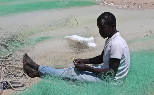 A fisherman mends his net on the beach at Gunjur fishing village. Image by Nosmot Gbadamosi. The Gambia, 2018.
