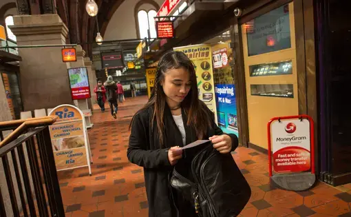 Au pair Lucila Espinosa Milay checks her receipt as she walks through a row of money transfer businesses after wiring part of her paycheck to siblings back in the Philippines, at Central Station in Copenhagen, Denmark, May 21, 2016. Milay pays the school fees for several of her eight siblings back home, sending money every 15 days. Remittances are a huge economic driver in the Philippines. Image by Allison Shelley. Philippines, 2016.