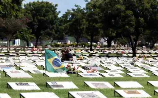 A cemetery field with rows of grave markers. Some of the graves have flowers. A Brazilian flag waves in the foreground. Two people visit a grave in the background.