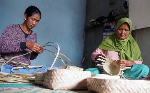 Two women are weaving baskets together.
