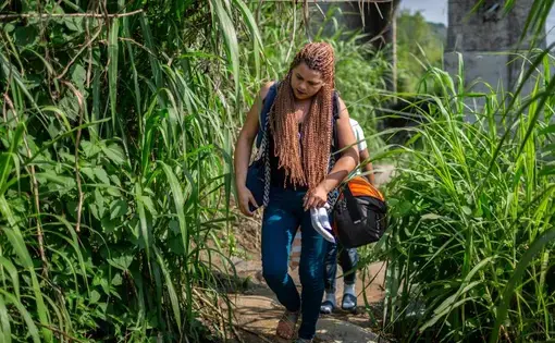 A young woman with long braids walks down a stone path with foliage on either side of her. She's wearing jeans and sandals and carries with her a duffel bag and a purse.