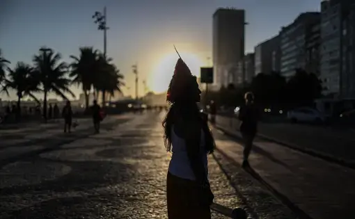 The silhouette of a woman wearing a headdress standing against the sun, as it sets in a city.