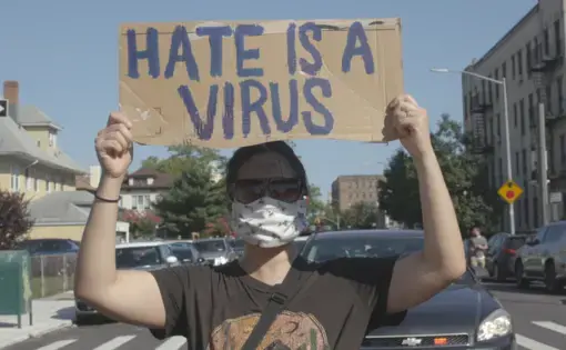 A masked person holds a sign that reads 'hate is a virus'