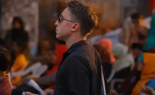 A man wears a black shirt and sunglasses. He carries a microphone. In the foreground, there are people sitting on plastic chairs.