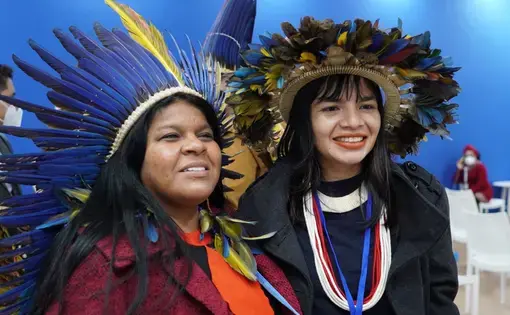 Two people pose for the photo. Both are wearing Indigenous clothing of orange and black colors.