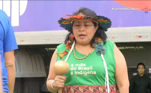 A person wears an Indigenous headdress and a green shirt while playing a small instrument.