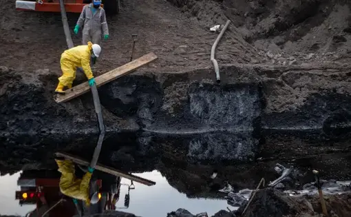 Two people in hazard suits are examining a polluted river full of oil.
