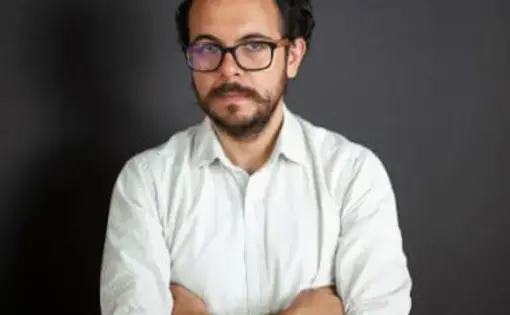 A man with short brown hair, black eyeglasses, a white shirt, and he crosses his arms.