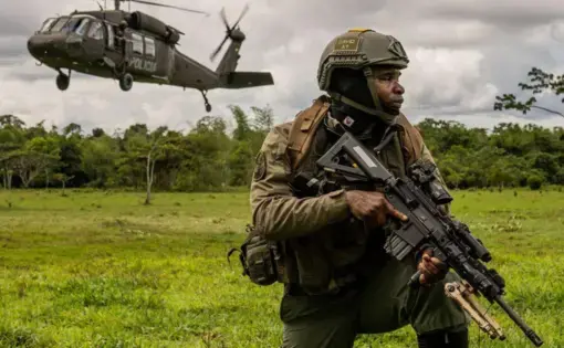 an armed soldier kneels in the middle of an open field while a helicopter flies away