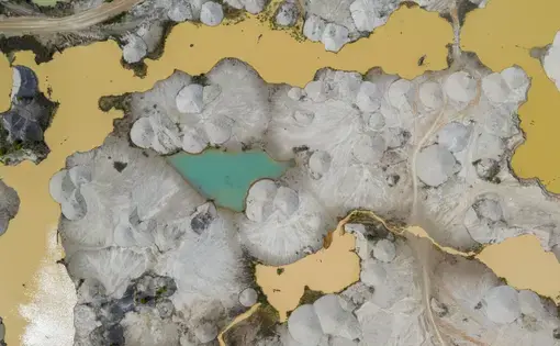 An aerial view of a centered, bright green-blue pond, surrounded by gravel and muddied waters.