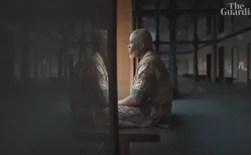 A Black man sits, his reflection appearing in the glass wall.