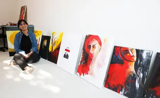 A woman sits on the floor next to a row of paintings that are on the floor propped up on a white wall