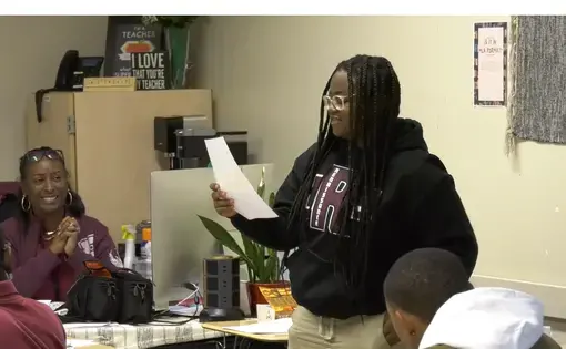 A student stands and reads an original blackout poem in class. A teacher looks on from their desk and smiles.
