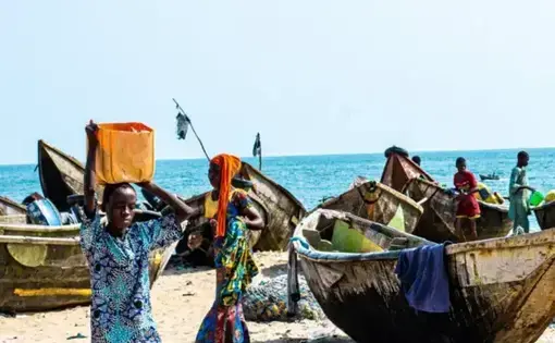 A young boy carries a bright orange plastic container on his head by the coast.