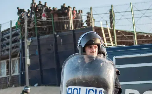 A border agent in riot gear stands in the foreground, with migrants behind a wire fence in the background