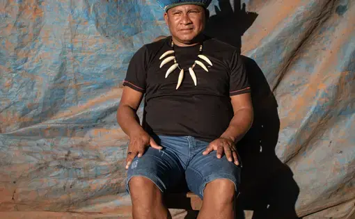 A man in black t-shirt sits atop a stool donning a traditional, elaborate hat.
