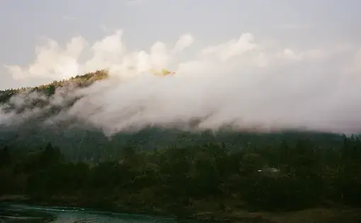 Clouds enshroud a green mountain, with a river at its base