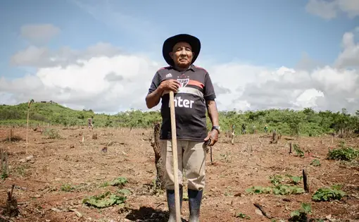 An olad man donning a hat stands in a field that he sows seeds in.