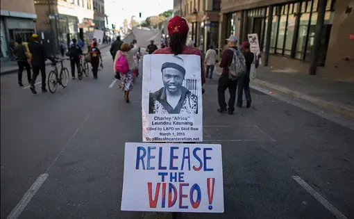 A man in the middle of a pedestrian street holds a sign reading "release the video"