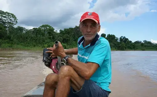 A man sits atop boat, sailing in a murky Amazonian river.