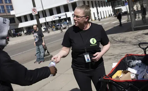 Community responder Kristina Paul, with Roots Wellness Center’s Appropriate Response Team, hands water to a passerby on Wednesday, March 25, 2026, in downtown St. Paul, Minn. The team handed out bus passes, Narcan and supplies to folks at a busy bus stop.