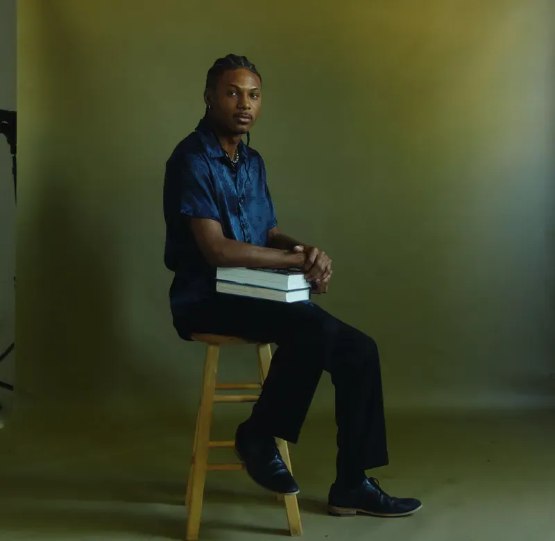 a Black man sits on a stool facing the camera holding textbooks on his knee