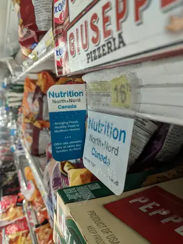 At a store in Pangnirtung, Nunavut, a sign for the Nutrition North program hangs above a frozen pizza (cost: $16.79). Inuit groups have been pressing Ottawa to reform the controversial subsidies for southern food flown into the North. Image by Julie De Meulemeester. Canada, 2018.&nbsp;