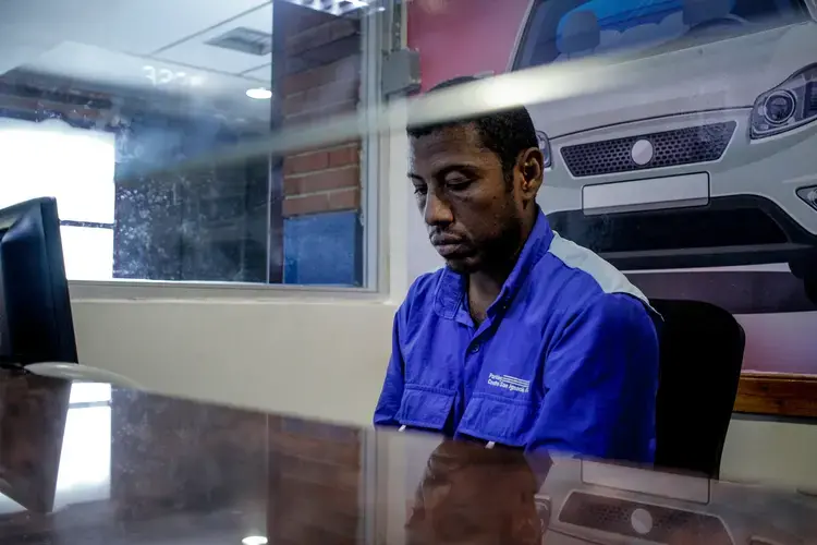 Carlos Román sits at his job post, his eyes staring down, wearing a bright royal blue uniform shirt. He works as a cashier in a parking lot&nbsp;at a shopping mall. He works the afternoon shift, from 2 p.m. to 7 p.m. It usually takes several hours to get back home to the satellite city of Guarenas because of the lack of public transportation. Image by Flaviana Sandoval. Venezuela, 2018.