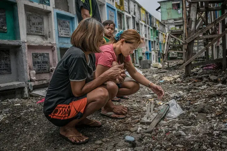 Mary Grace Llimit, in the pink shirt, at Novatas Catholic Cemetery. Her 16-year-old daughter, Liezel, was killed alongside her boyfriend. Liezel’s autopsy showed she was three months pregnant. Mary Grace was working in Singapore when she received the news of her daughter’s death. Liezel was shot eight times, including four bullets&nbsp;to the head, leaving her unrecognizable, says her mother. With Mary Grace are her mother, Appolinaria Salita, and her youngest son, Noel, who is three years old. Image by James Whitlow Delano. Philippines, 2017.