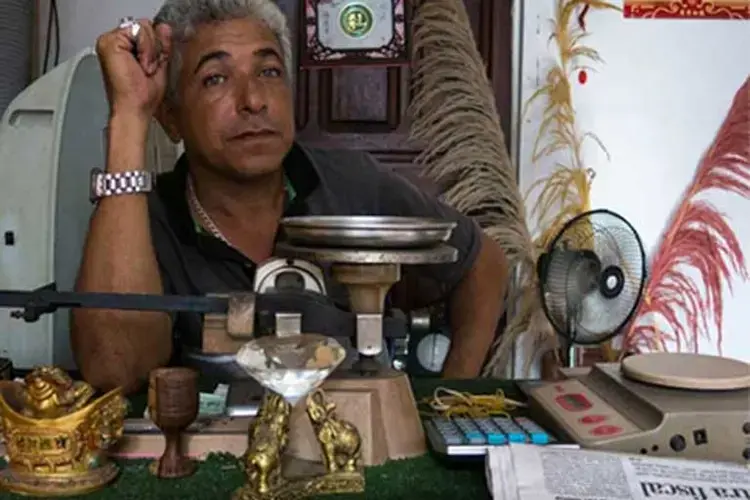 A Tumeremo gold pawn shop owner sits behind his desk while enjoying the luxury of a cooling desk fan. Image by Bram Ebus. Venezuela, 2017.<br />
