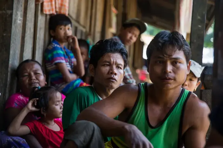 Workers and their families, including Zaw Myo Htike (foreground in green sleeveless shirt), gather on their porches at a small village where migrant workers for Supowin Palm Oil live outside Kawthaung, Myanmar. Many workers on this plantation, some of which have been here for over a decade, want to return home. With their low wages, however, few can afford the trip for their entire family, and they feel stuck. Image by Taylor Weidman/PRI. Myanmar, 2016. 