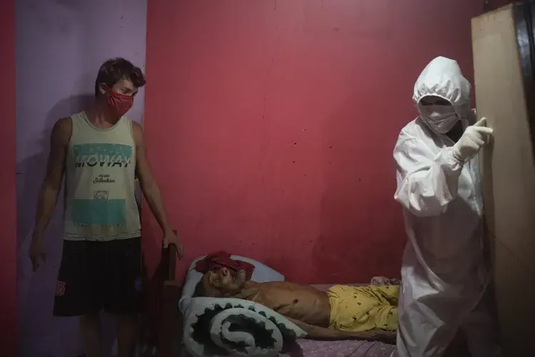 A worker from the SOS Funeral service, wearing protective gear amid the new coronavirus pandemic, is helped by caregiver Leandro Beiton as he prepares to remove the body of Edgar Silva in Manaus, Brazil, Tuesday, May 12, 2020. Image by Felipe Dana / AP Photo. Brazil, 2020.