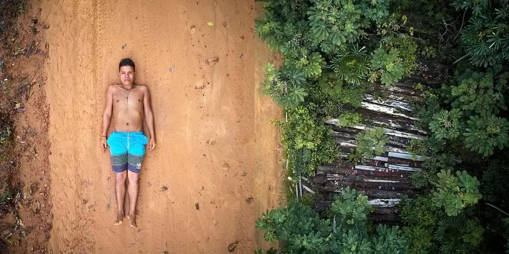 Ednei, 20, is a young Arapiun indigenous leader who has recently joined the land guard team at the Indigenous Territory of Maro (TI Maro) in the Maro River. The team carries out regular surveillance beats across the rainforest, watching for illegal loggers and poachers stealing from their sacred land. TI Maro covers some 42,000 hectares of untouched, pristine rainforest. Under the strong leadership of chief Dada Borari, the community has been fiercely fighting to defend themselves from illegal loggers who extract wood from their ancestral land. Since their territory was officially recognized, they have been keeping a close watch. Right: 26 huge precious logs captured by the TI Maro team in one of their beats, now lying by the road track. Measuring 1 to 2 meters in diameter, the 26 logs rot to fertilize the land. Left: Ednei is portrayed lying on the road tracks left by logger trucks driving close to the borders of the Indigenous Maro Territory. Image by Pablo Albarenga. Brazil, 2019.