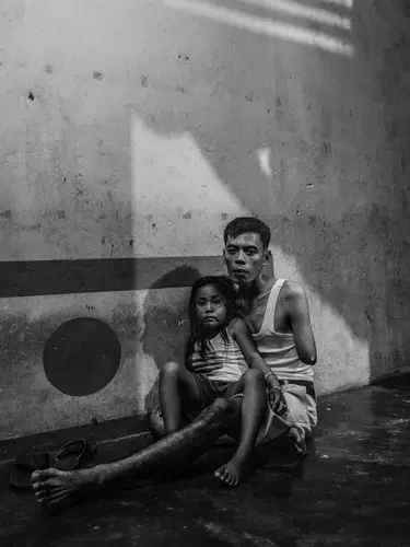 Misnan sits with his daughter Delisa in their home in Kandis. He lost his arm when he crashed his motorbike while carrying a scythe. He was on his way to work at a plantation. Image by Xyza Cruz Bacani. Indonesia, 2018.