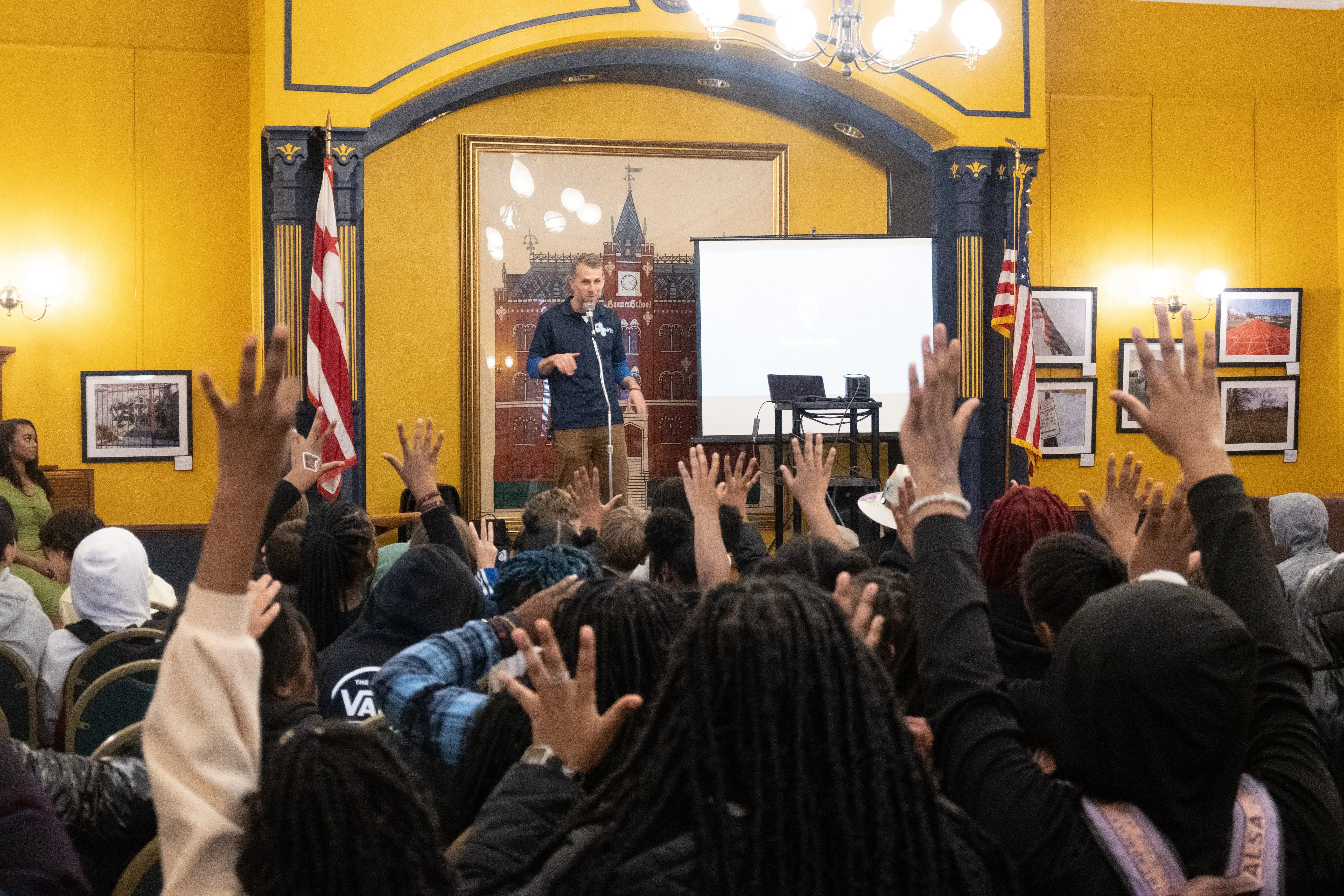 A man stands on stage at the Charles Sumner gallery holding a microphone and the audience of students raise their hands
