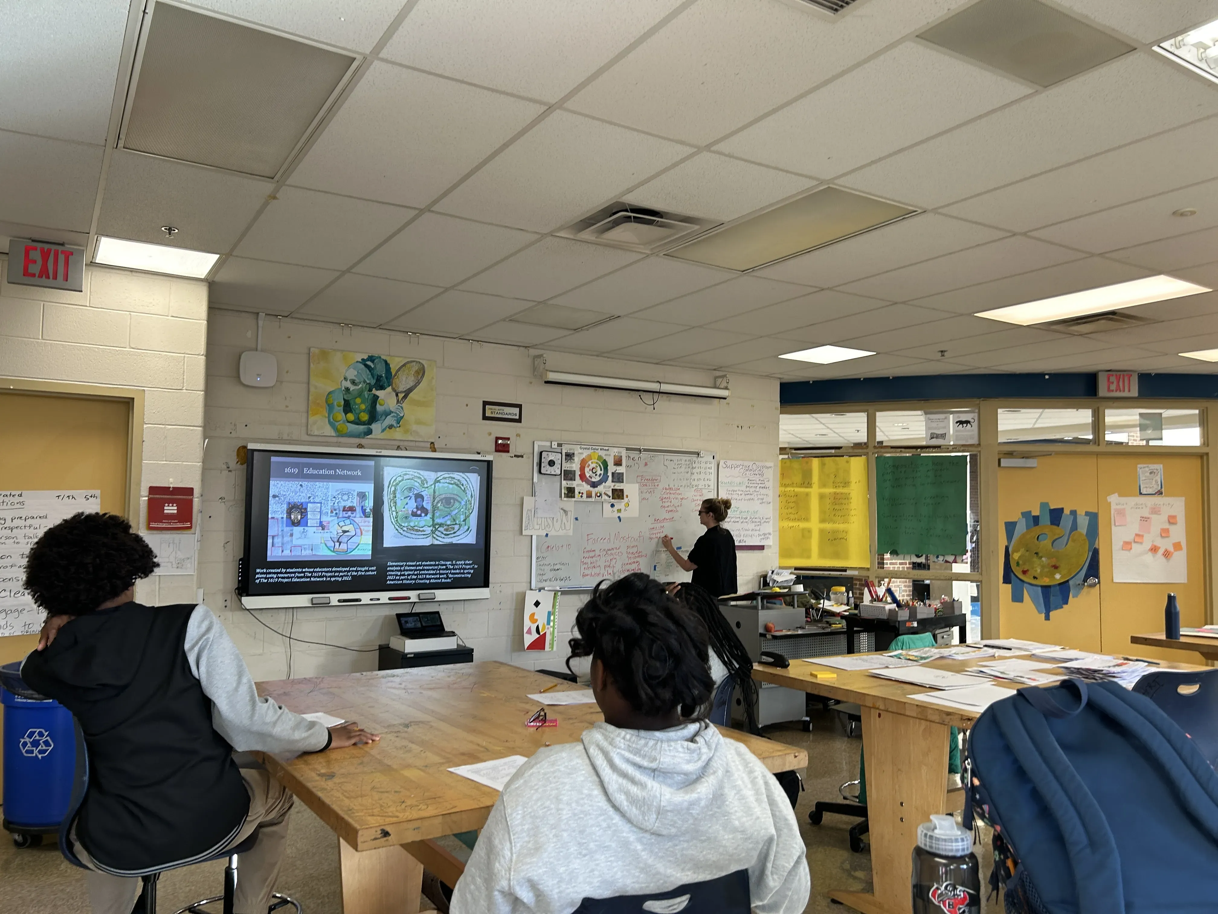 Students observe a teacher who writes at a whiteboard. There is a projection of student artwork on a screen.