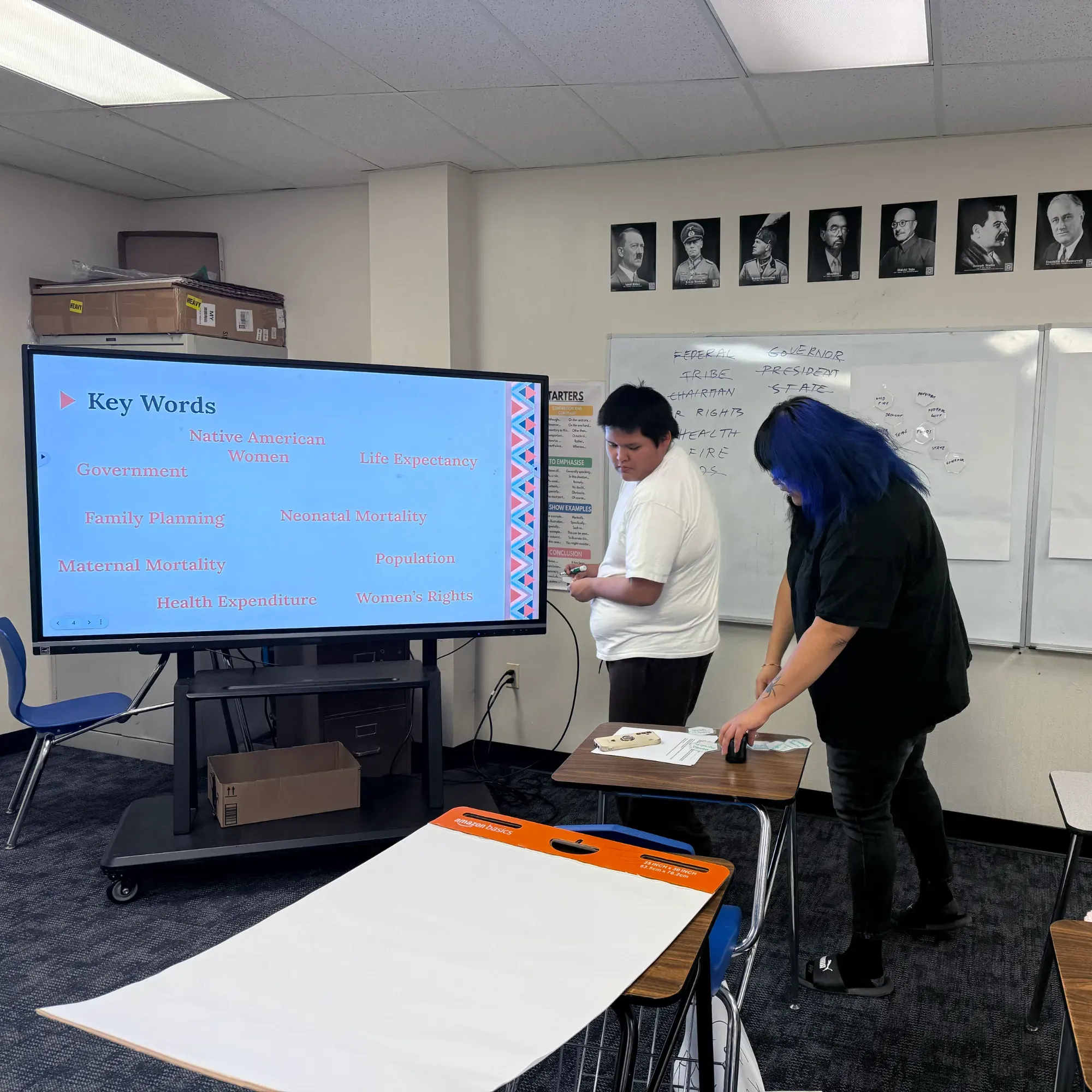 Two students review their notes in front of the classroom smartboard