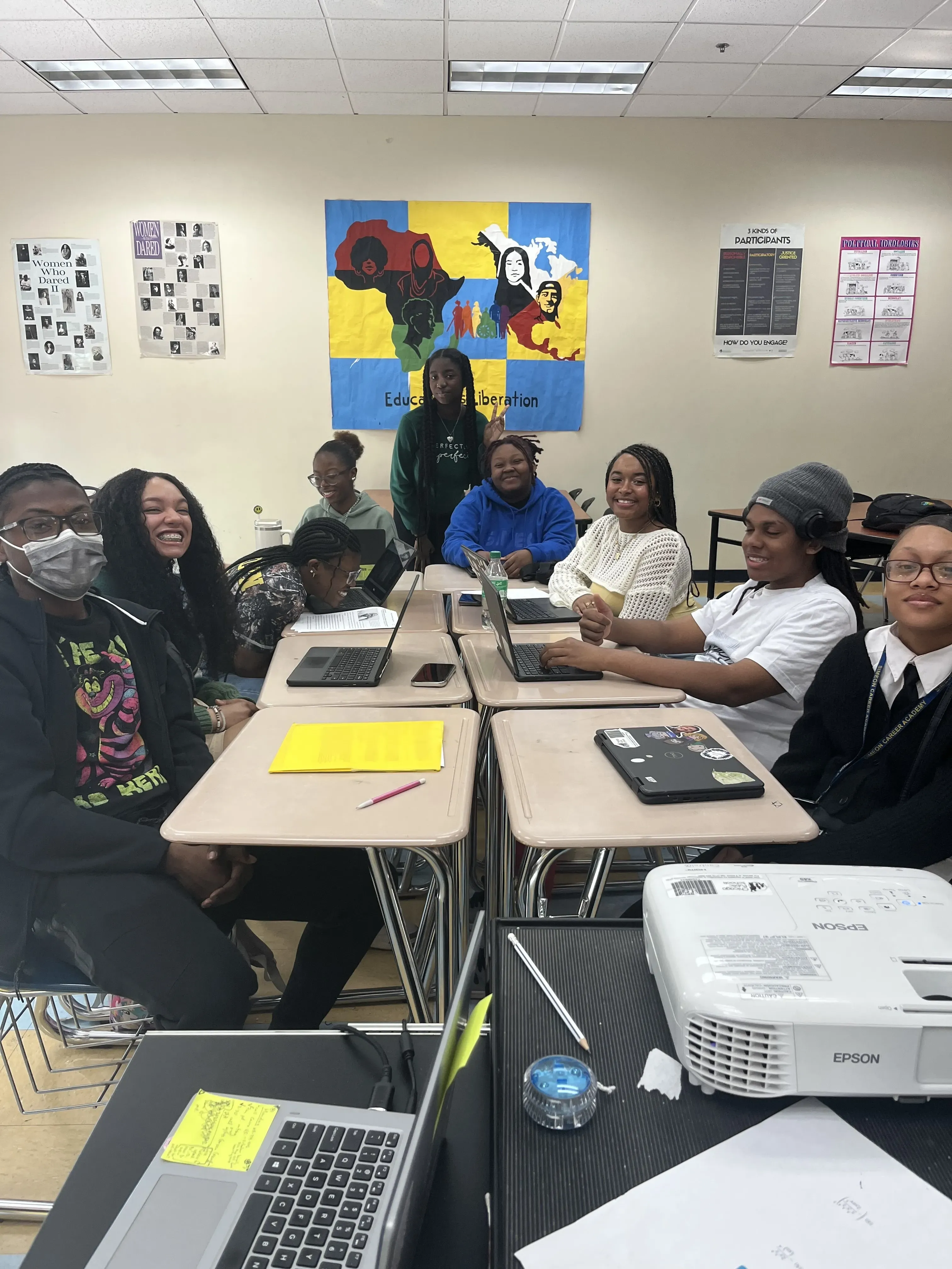 A group of high school students sit at desks and smile for the camera.