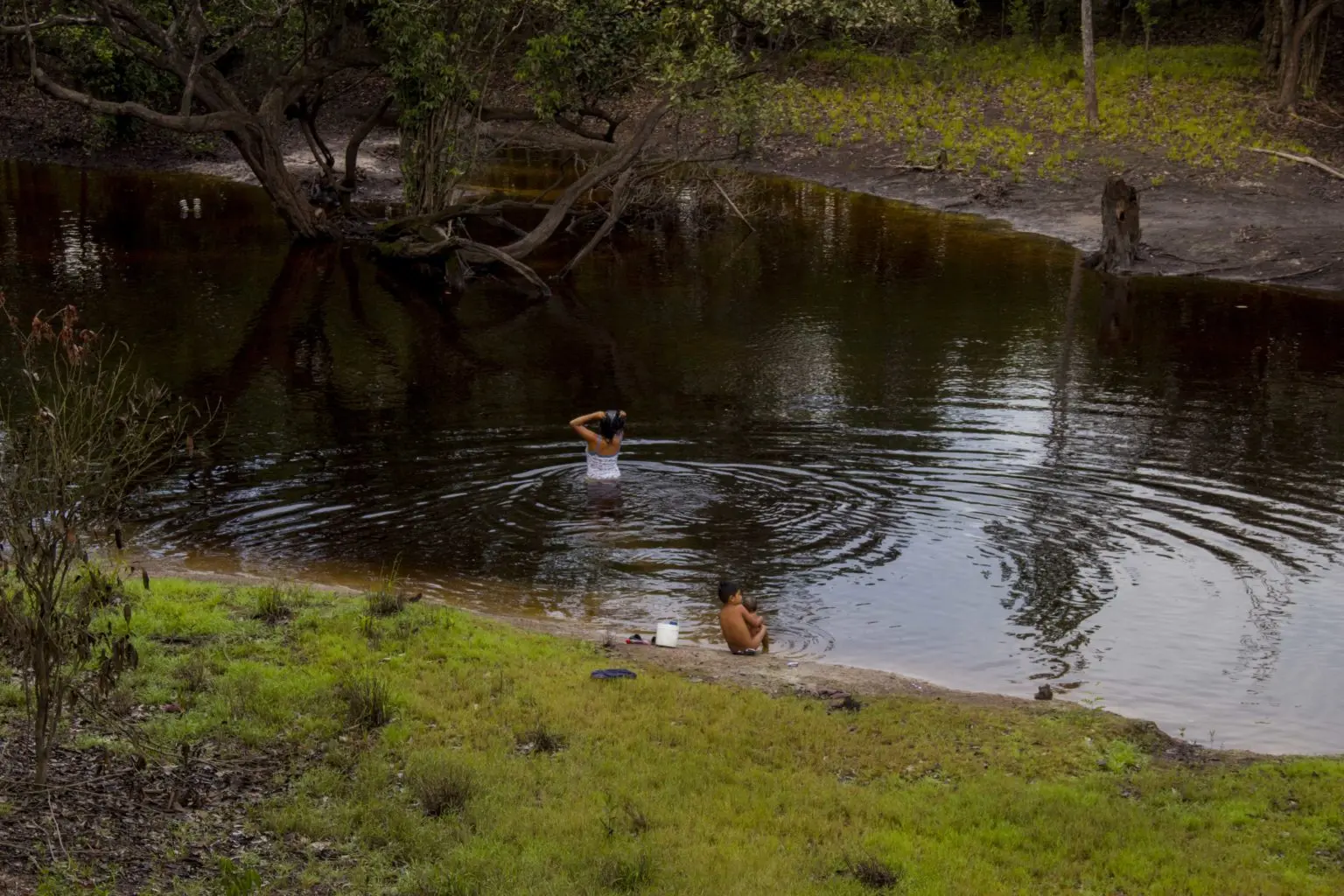 una señora se baña en las aguas negras de un rio. Su hijo desnudo amarca a un bebé en la orilla 