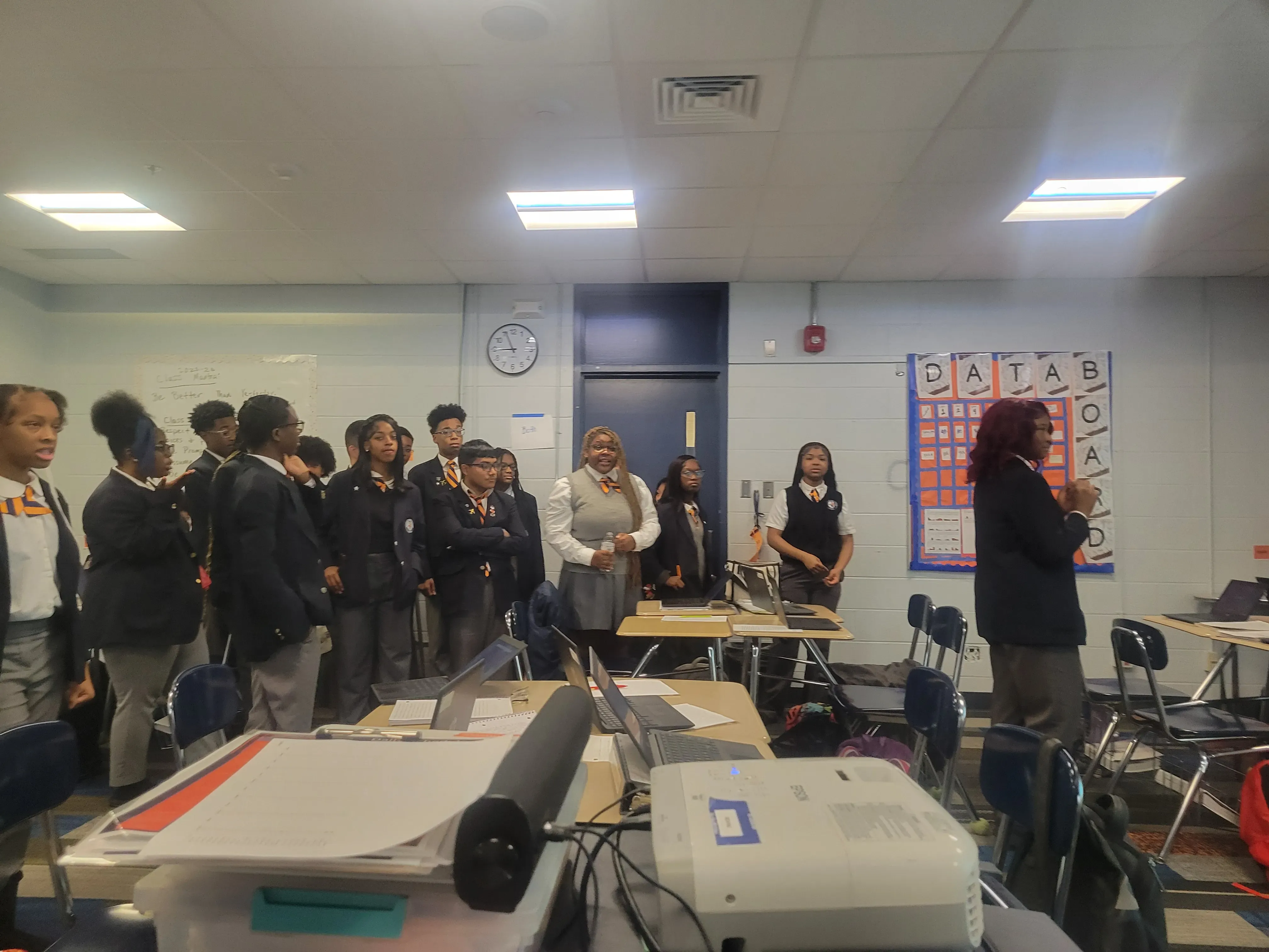 Students in a classroom stand together while a peer presents their project