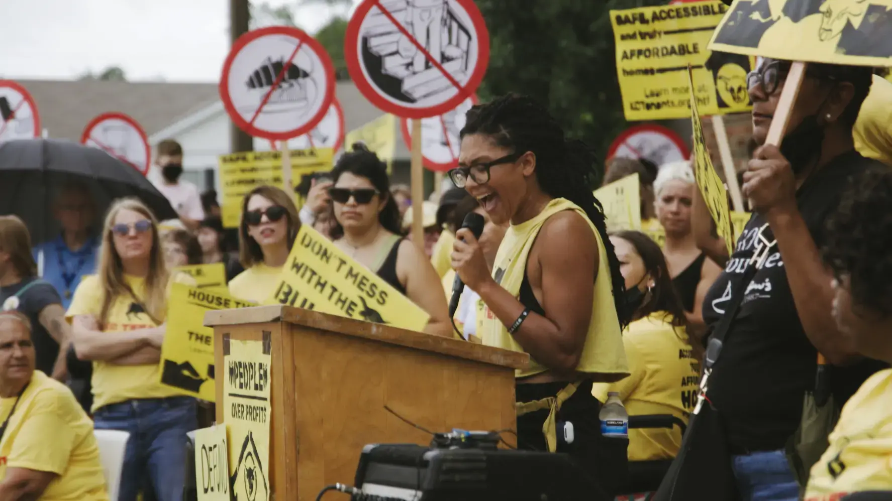 A woman in yellow speaks at a podium at a protest, surrounded by other protestors. The protestors are carrying yellow signs that say