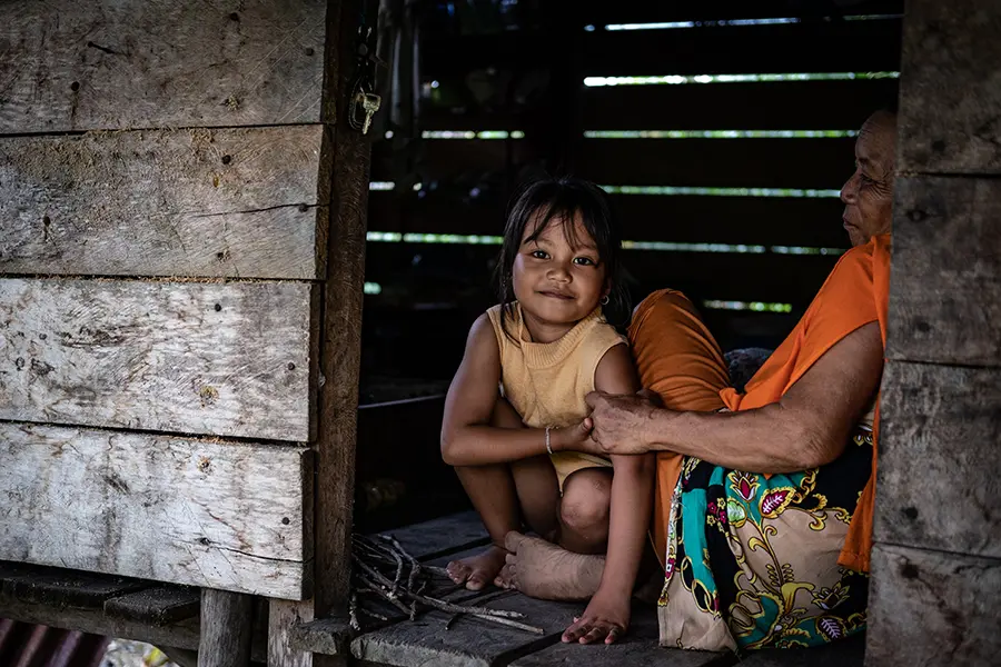 a young girl smiles at the camera. She sits next to an elder. They are both sitting in their wooden house.