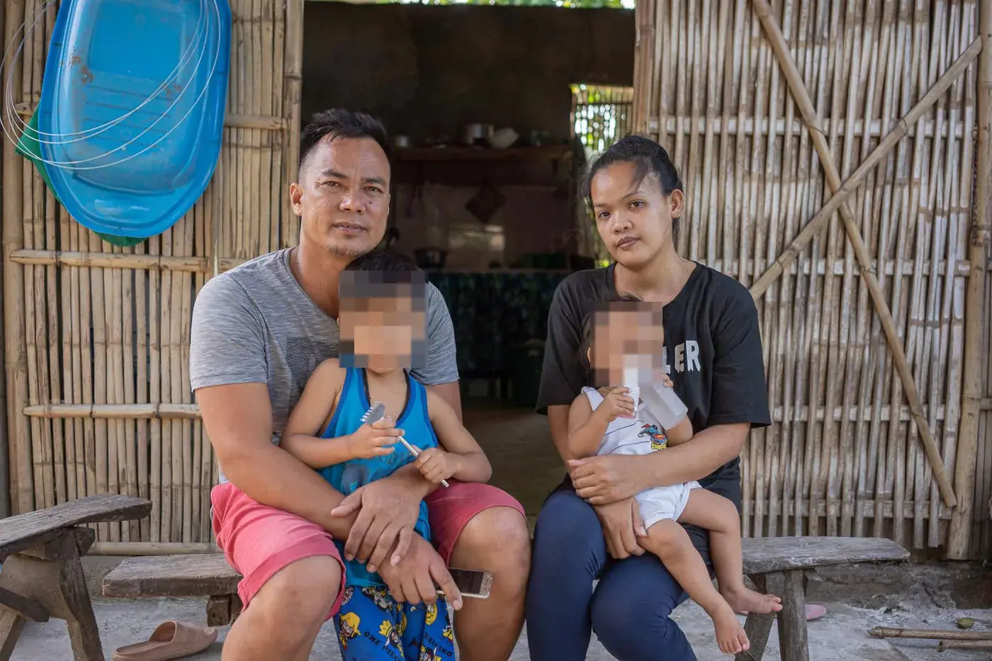 A couple sits with their two children outside of their home; the children's faces are pixelated