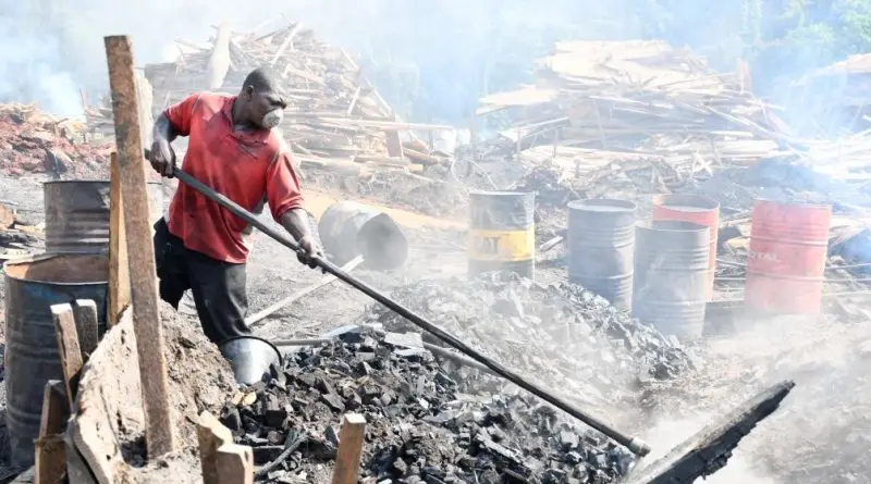 A worker shovels charcoal into a pile. He wears a mask. In the background are piles of wood scraps and metal containers. 