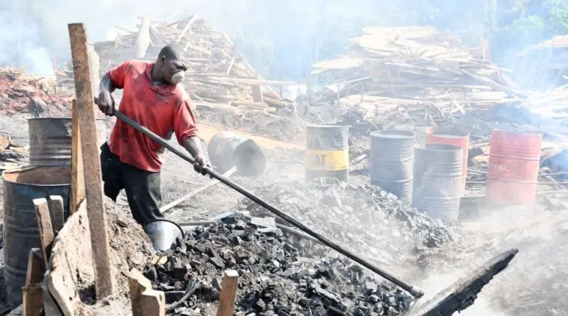 A worker shovels charcoal into a pile. He wears a mask. In the background are piles of wood scraps and metal containers.