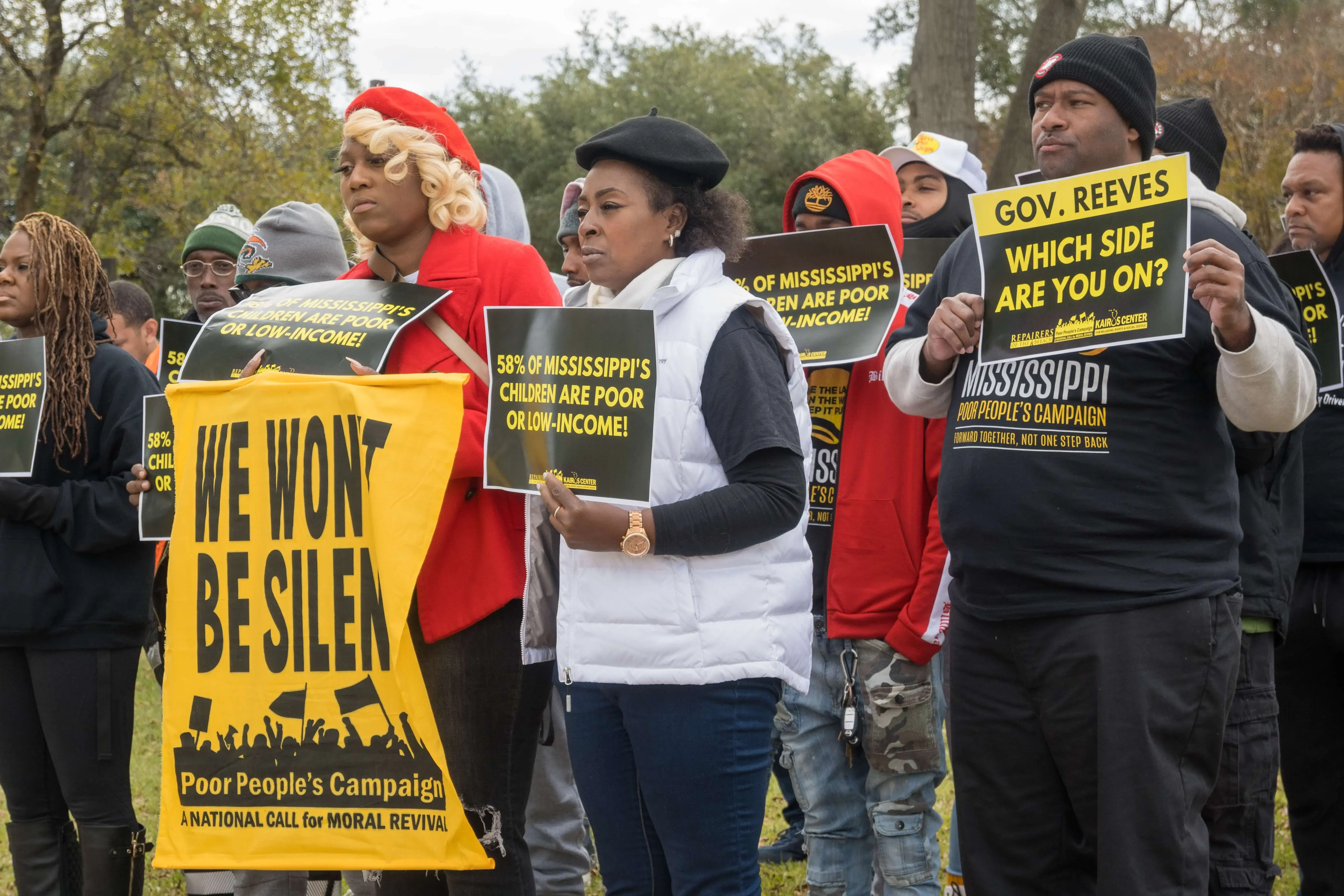 Protestors stand in a crowd holding signs saying, "We won't be silent" and "58% of Mississippi's children are poor or low-income!"