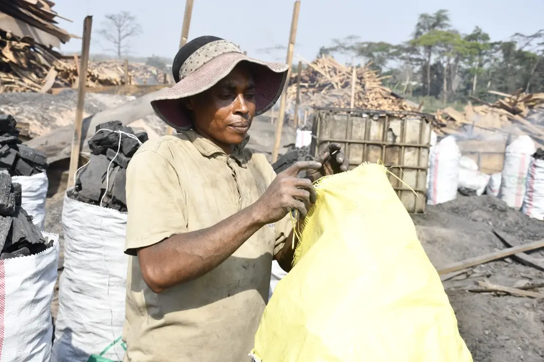 A worker prepares a bag used for packaging charcoal. Scraps of wood are piled up in the background. There are multiple similar bags filled with charcoal.