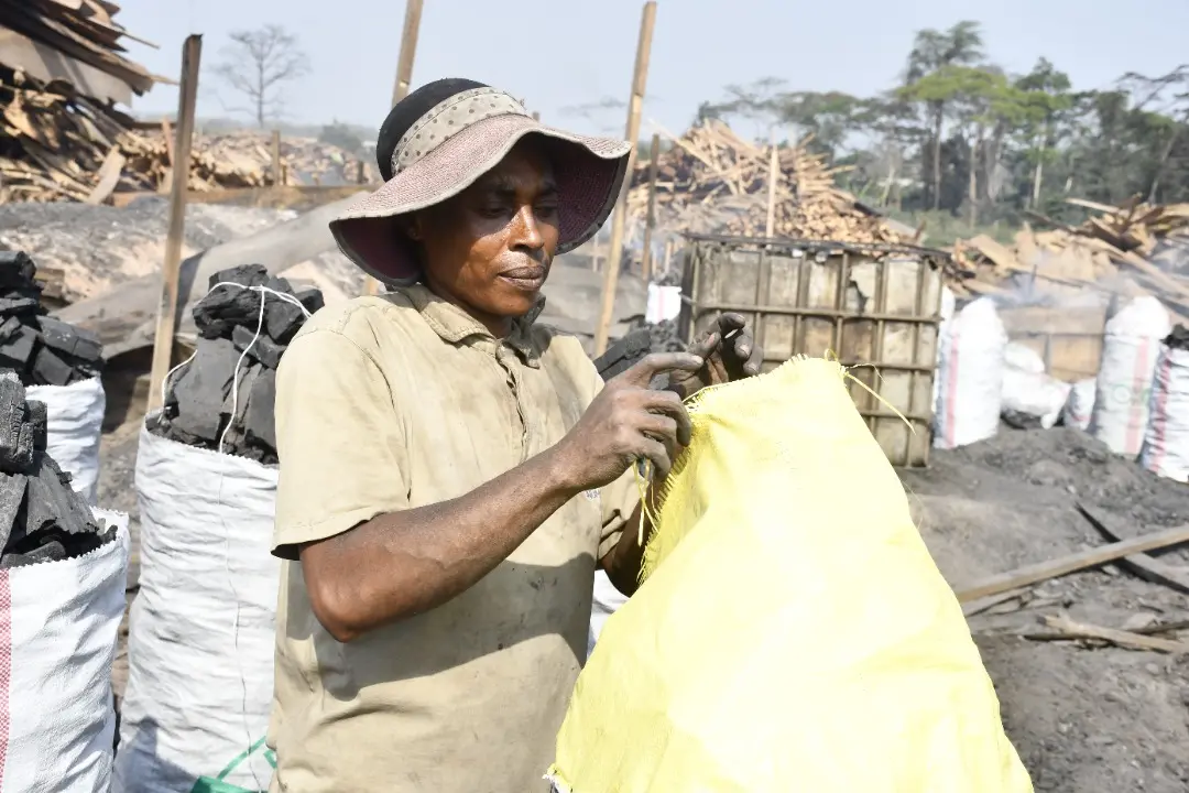 A worker prepares a bag used for packaging charcoal. Scraps of wood are piled up in the background. There are multiple similar bags filled with charcoal. 
